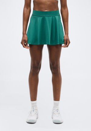 Femme portant une jupe de sport verte, des chaussettes blanches et des baskets blanches, debout devant un fond clair uni.