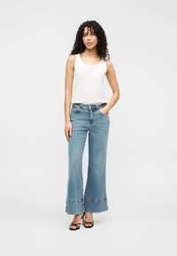 Τζιν παντελόνι σε ανοιχτό μπλε με φαρδιά πατζάκια και ακατέργαστα τελειώματα, με κλασικό σχέδιο πέντε τσεπών και επίχρυσο κούμπωμα. Συνδυασμένο με εφαρμοστή λευκή φανέλα χωρίς μανίκια.