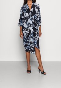 Kvinna som bär en svartvit abstrakt blommig midiklänning med V-ringning och draperad front, matchad med svarta klackade muletofflor.