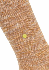 Calzino a maglia arancione e bianco mélange con piccolo emblema circolare giallo vicino alla caviglia su sfondo bianco.