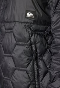 Sort pufferjakke med hexagonal quiltet mønster, der har en lynlåslomme og Quiksilver-logo patch på brystet. Glat, skinnende stof.