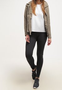 Olivově zelená bunda na zip, bílý svetr, černé legíny s logem a černé sportovní boty. Neformální outfit se vrstvami textur.