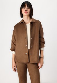Giacca camicia in suede marrone con colletto, bottoni a scatto e vestibilità leggermente oversize. Indossata sopra un maglione di un leggero colore crema.