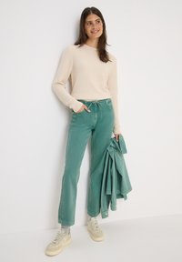 Suéter de punto beige claro, pantalones vaqueros verdes de pernera ancha con cintura ajustable y zapatillas beige, sosteniendo una chaqueta verde a juego.