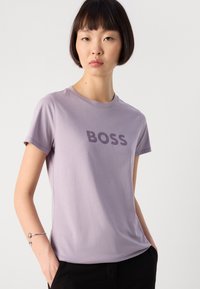 Jauna moteris su trumpais tamsiais plaukais, vilkinti šviesiai purpurinę marškinėlius su užrašu "BOSS" ir juodas kelnes, pozuojanti su rankomis kišenėse.