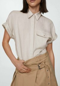 Beige linen shirt with short rolled sleeves, button closure, and a single chest pocket. Worn with a beige high-waisted skirt featuring a belt.