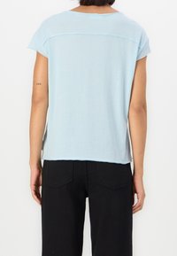 Světle modrý top s krátkým rukávem vyrobený z měkké látky, s jemným horizontálním švem na zadní straně a uvolněným střihem. Nosený s černými kalhotami.