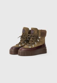 Botas de ante marrón con un puño de piel de pelaje marrón texturizado, punta redonda y suela de plataforma gruesa. Presenta ojales de metal y acentos de cuero.
