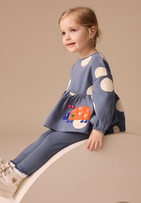 Sinine täpiline topp, millel on värviline sisalikud tikand ja kogutud vöökoht, koos ribakujuliste siniste retuusidega ja valgete tossudega.