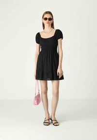 Γυναίκα ντυμένη με μαύρο κοντό φόρεμα με φουσκωτά μανίκια, μαύρες σαγιονάρες, γυαλιά ηλίου, κολιέ από πέρλες και κρατώντας μια μικρή ροζ τσάντα.