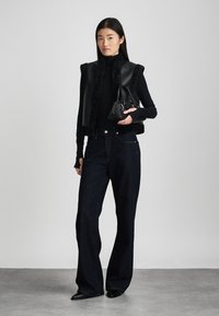 Μαύρο γιλέκο από τεχνητό δέρμα με γούνα στο περιθώριο, μαύρος ψηλός λαιμός, σκούρες φαρδιές παντελόνες και μαύρη τσάντα με διακριτικές μεταλλικές λεπτομέρειες.