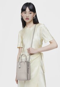 Borsa a tracolla compatta di colore grigio chiaro con due manici e una tracolla staccabile. Presenta il logo del marchio impresso e un ciondolo decorativo. Texture liscia.