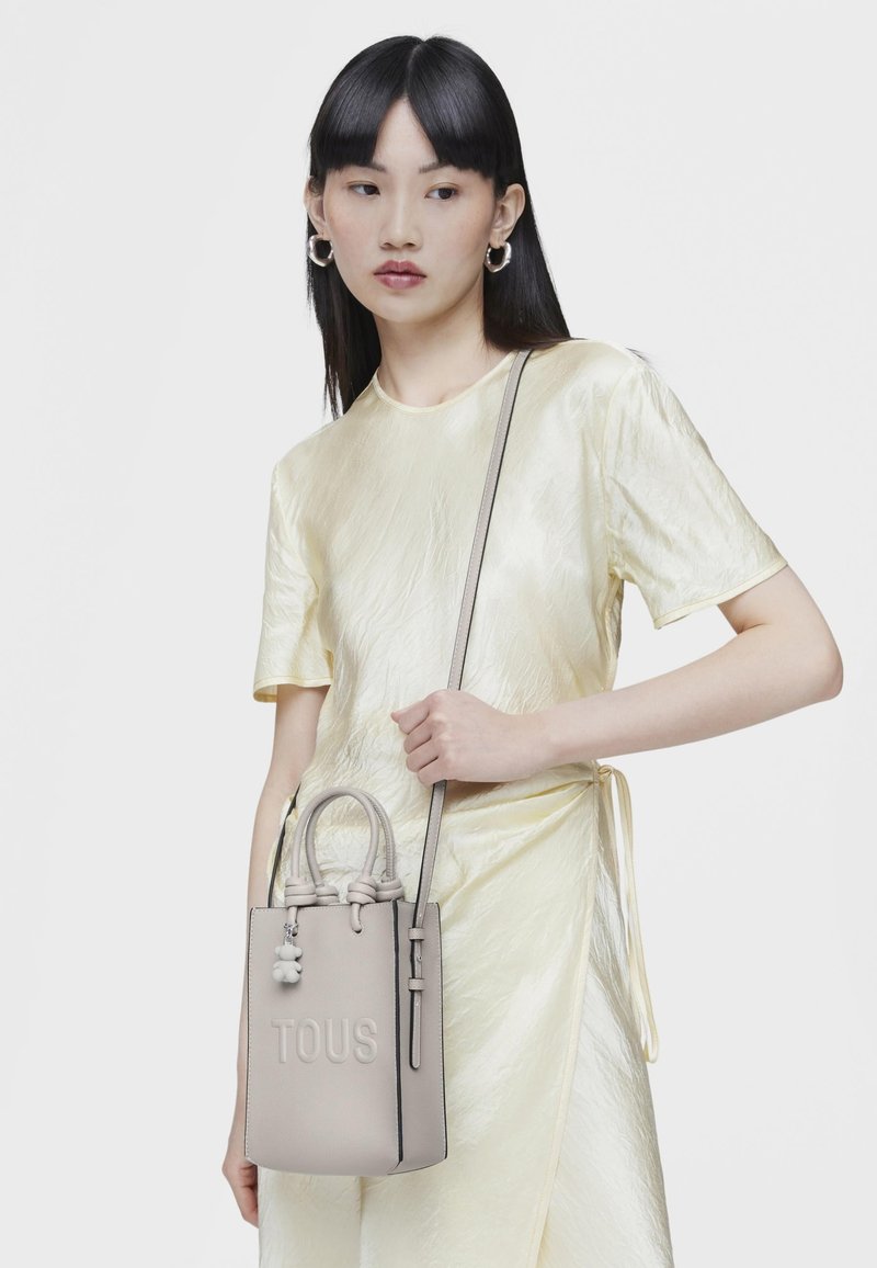 Borsa a tracolla compatta di colore grigio chiaro con due manici e una tracolla staccabile. Presenta il logo del marchio impresso e un ciondolo decorativo. Texture liscia.