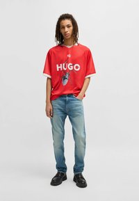 Maglietta rossa a maniche corte con dettagli bianchi e stampa "HUGO" che presenta un grafico di ragno, abbinata a jeans blu chiaro e scarpe nere.