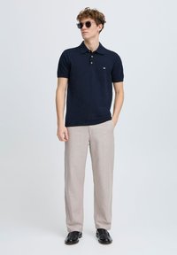 Polo cu mânecă scurtă de culoare bleumarin, cu guler și nasturi aurii, asortat cu pantaloni bej deschis și pantofi negri, evidențiind un design ajustat.