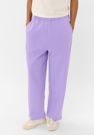 Personne portant un pantalon de survêtement violet clair avec des poches et des baskets beige, debout devant un fond blanc uni.