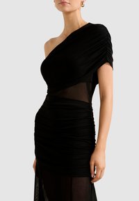 Sieviete valkā melnu asimetrisku piegulošu kleitu ar ručingiem un caurspīdīgiem tilla paneļiem, papildinātu ar zelta lokveida auskariem.