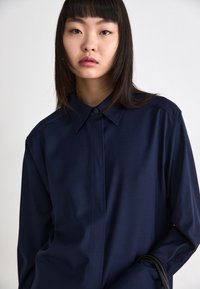 Mørkeblå skjorte med knapper, strukturert krage, lange ermer og en glatt tekstur. Har frontstolpe og minimalistisk design.