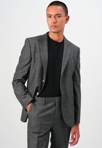 Grau strukturierte Sakkojacke mit zwei Knöpfen, passenden Hosen und einem schwarzen T-Shirt darunter, getragen von einem Model vor einem neutralen Hintergrund.