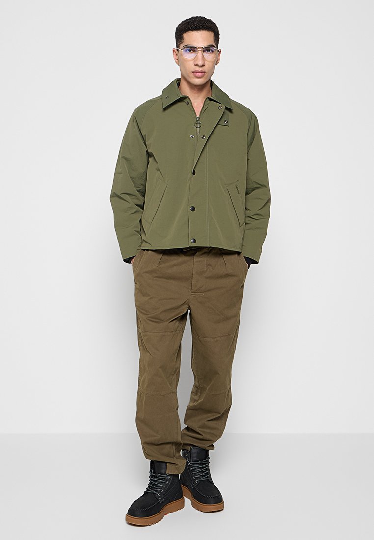 Olijfgroene jas met een opgerolde kraag en knopen aan de voorkant, gecombineerd met losse bruine broek en zwarte laarzen, staand tegen een effen achtergrond.