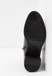 Semelle d'une chaussure en cuir marron montrant un talon texturé et des motifs répétitifs embossés "ANNA FIELD" avec le logo sur une surface blanche propre.