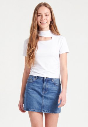 Jeune femme aux longs cheveux portant un haut blanc à manches courtes avec une découpe au niveau de la poitrine et une mini-jupe en jean bleue, souriante et debout.