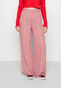 Calças largas em tecido vermelho claro com padrão floral branco. Cintura elástica e textura fluida, combinadas com sapatos casuais em cor bordô.