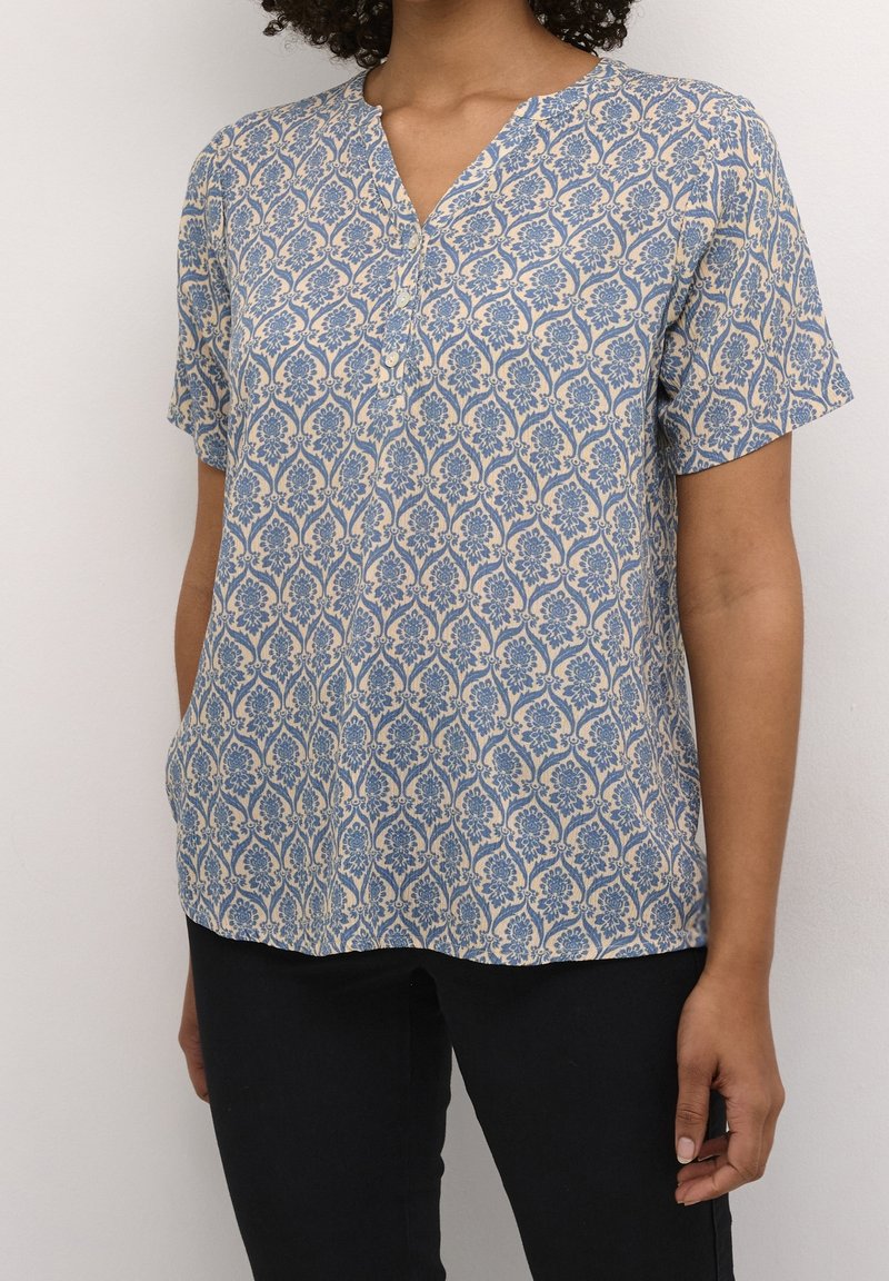 Kortærmet bluse i let stof, med et blåt blomster mønster på en beige baggrund, V-hals og knapdetalje ved kraven.