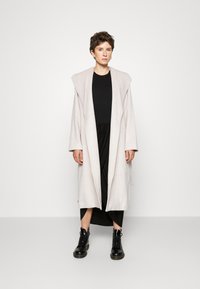 Abrigo largo de lana beige con solapas anchas, frente abierto y corte recto. Llevado sobre un vestido negro, combinado con botas negras con cordones.