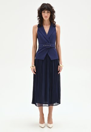 Femme aux cheveux bouclés foncés portant un top blazer bleu marine sans manches ceinturé et une jupe midi plissée assortie, des talons beiges et des bracelets dorés.