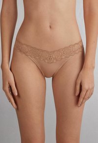 Nude kantenv thong met een bloemenpatroon langs de tailleband, voorzien van delicate golvende randen en een gladde stof.