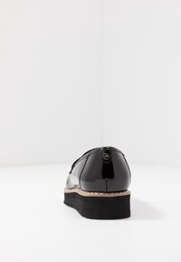 Svart lackskinnssko med rundad bakdel, strukturerad sula och en liten logotypdetalj på hälen. Slät yta med platt design.