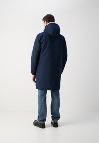 Abrigo largo azul marino con capucha, que presenta una textura suave y un cuello forrado. Combinado con jeans azul claro y zapatos negros.