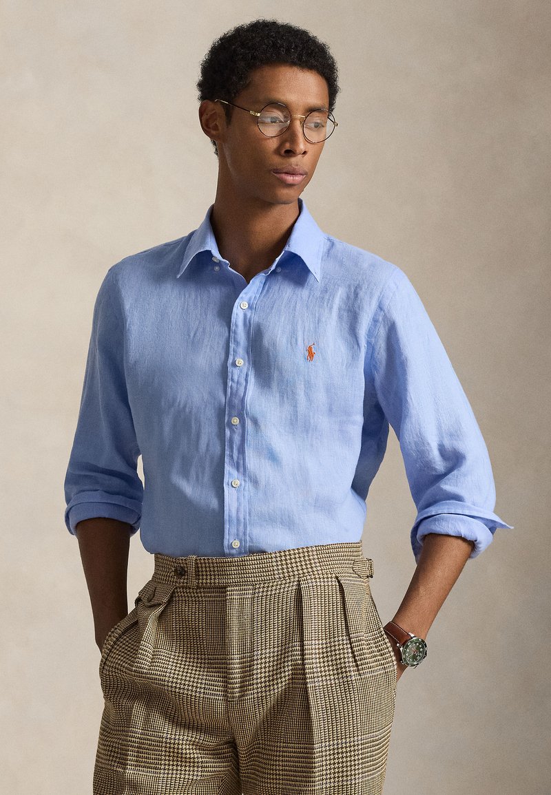 Ανοιχτό γαλάζιο λινό πουκάμισο με γιακά με κουμπιά και πορτοκαλί λογότυπο, συνδυασμένο με ψηλόμεσο καρό παντελόνι σε καφέ χρώμα. Διαθέτει μανίκια γυρισμένα και ρολόι.