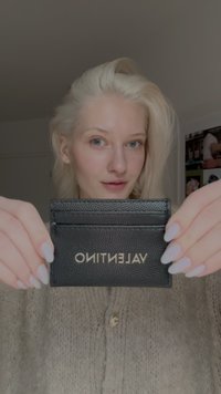 Cartão de couro texturado preto com "VALENTINO" em letras douradas; segurado com duas mãos, posicionado contra um fundo neutro.