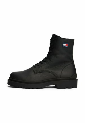 Botas de cuero negro con cordones, superficie texturizada, suela de goma resistente y un pequeño logo en rojo, blanco y azul en el lateral.