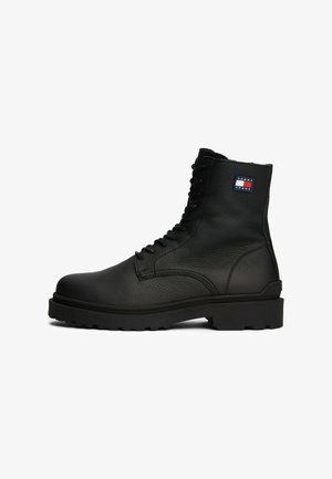 Bottes en cuir noir à lacets avec surface texturée, semelle en caoutchouc robuste, et un petit logo rouge, blanc et bleu sur le côté.