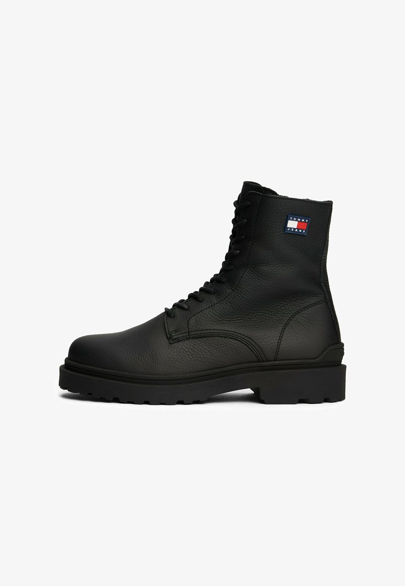 Botas de cuero negro con cordones, superficie texturizada, suela de goma resistente y un pequeño logo en rojo, blanco y azul en el lateral.