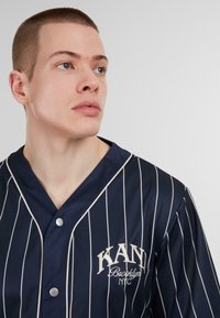 Camisa de punto de rayas marinas con un acabado texturizado. Presenta finas rayas blancas y un logo bordado "KANI Brooklyn NYC".