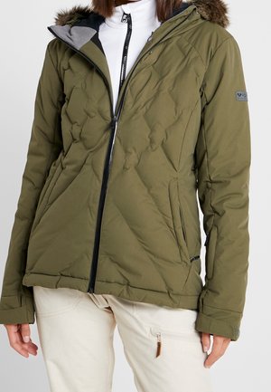 Personne portant une veste matelassée vert olive avec capuche bordée de fourrure, fermeture éclair noire et pantalon beige avec poche zippée.