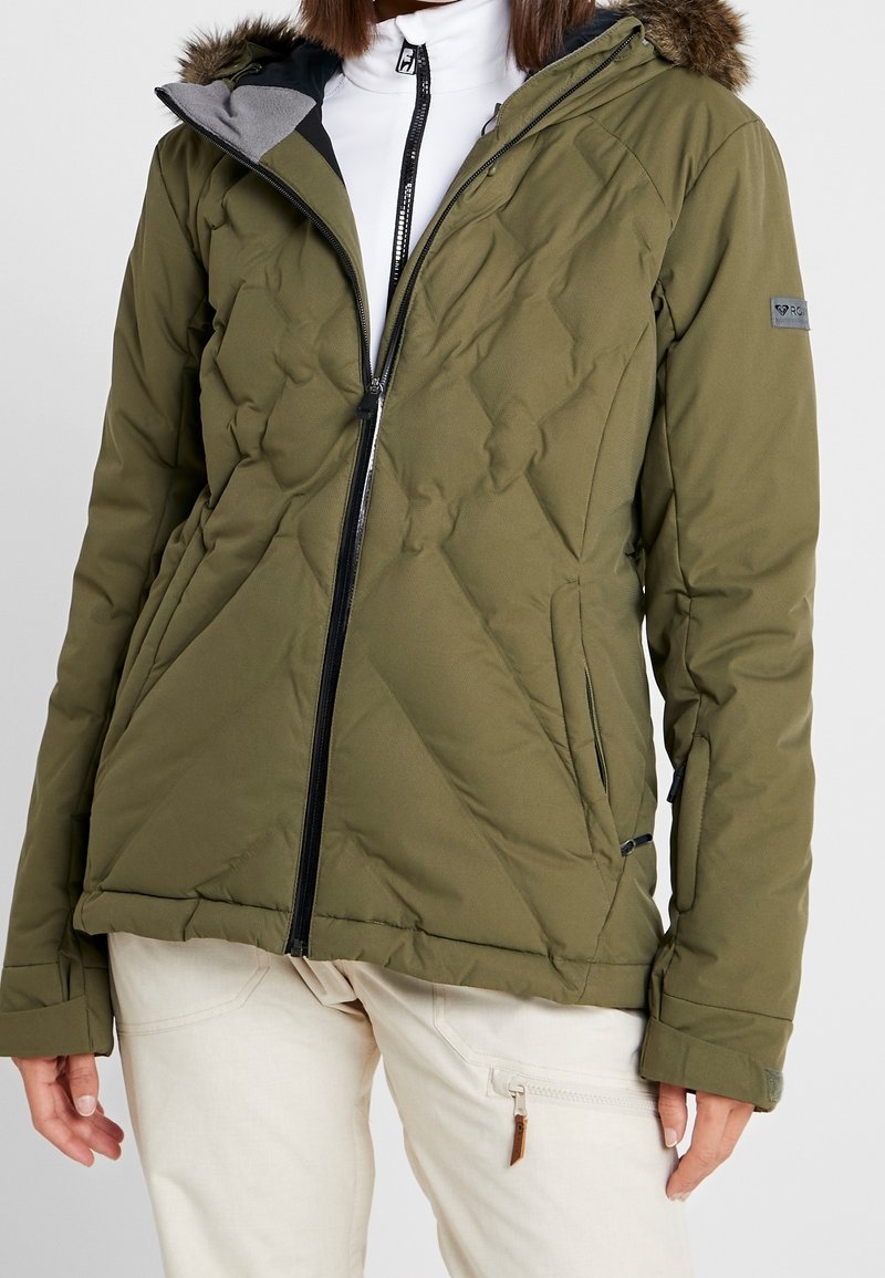 Personne portant une veste matelassée vert olive avec capuche bordée de fourrure, fermeture éclair noire et pantalon beige avec poche zippée.