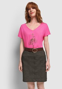 Mujer llevando una camiseta rosa de cuello en V con un diseño estilizado de flamenco y una falda oscura con botones de color gris y un cinturón marrón.