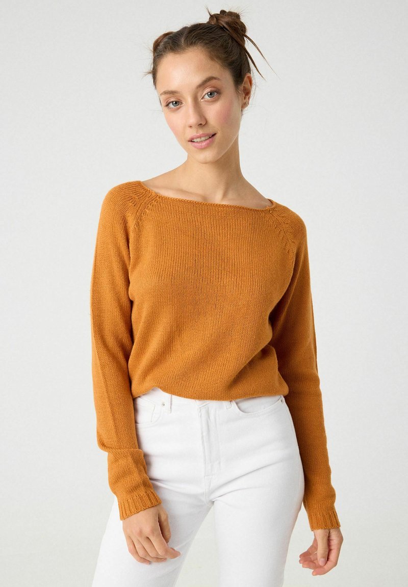 Suéter de color óxido con cuello barco, puños acanalados y un corte relajado. Llevado sobre unos pantalones vaqueros blancos de cintura alta, con una textura suave.