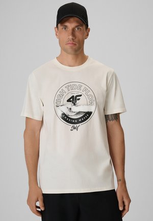 Mężczyzna w czarnej czapce i białej koszulce z napisem „High Tide Flow, Chasing Waves, Surf” oraz grafiką fal i deski surfingowej na klatce piersiowej.