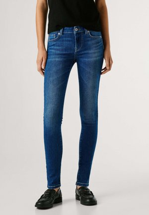 Person, die blaue Slim-Fit-Jeans, ein schwarzes Oberteil und schwarze Loafers trägt, steht vor einem schlichten hellen Hintergrund.