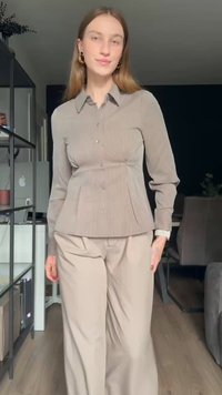 Young woman with long hair wears beige button-up shirt and matching wide-leg pants, standing in a sunlit modern living room.