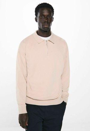 Langarm-Pullover in hellbeige mit Kragen und halbem Reißverschluss. Weicher Stoff, lässige Passform und gerippte Bündchen. Wird über einem weißen T-Shirt getragen.