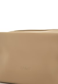 Beige vanntett bag med preget "RAINS"-logo. Glatt tekstur og rektangulær form med glidelås på toppen, minimalistisk design.