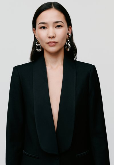 Black blazer with a deep V-neckline, satin lapels, and side pockets. Model wears large, abstract silver earrings. Smooth fabric texture.