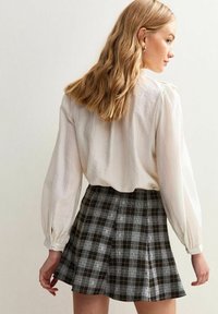 Young woman with wavy blonde hair wearing a white long-sleeve blouse and a black-and-white checkered skirt, facing sideways.